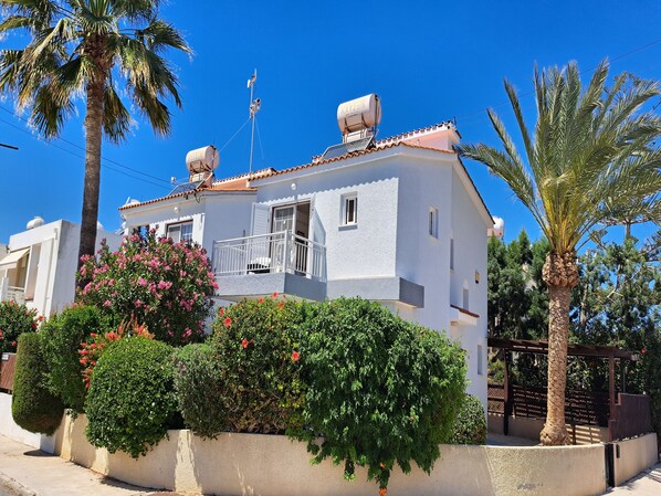 A white building with a balcony, surrounded by greenery and palm trees.