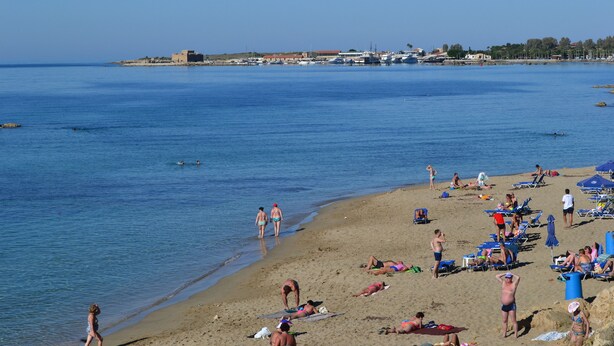 Een strand met mensen die zonnebaden en zwemmen, een heldere lucht en gebouwen op de achtergrond.