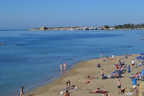 A beach with people sunbathing and swimming, a clear sky, and buildings in the background.