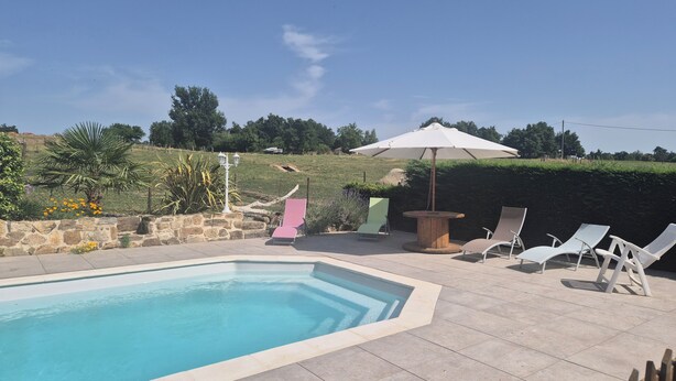 Une piscine entourée d’un mur de pierre et de verdure, avec des chaises longues et un parasol.