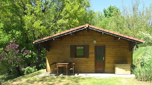 Una cabaña de madera con techo de tejas, una puerta principal y un pequeño porche con dos sillas y una mesa de madera.