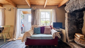 A cozy room with a wooden ceiling, a stone fireplace, a sofa with cushions, a television, and a window with curtains. - Remote Cottage with Woodburner Surrounded by Farmland  (Redruth)