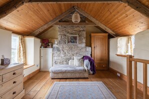 A cozy attic bedroom with a stone wall, wooden ceiling beams, a bed, a dresser, and a window with curtains.