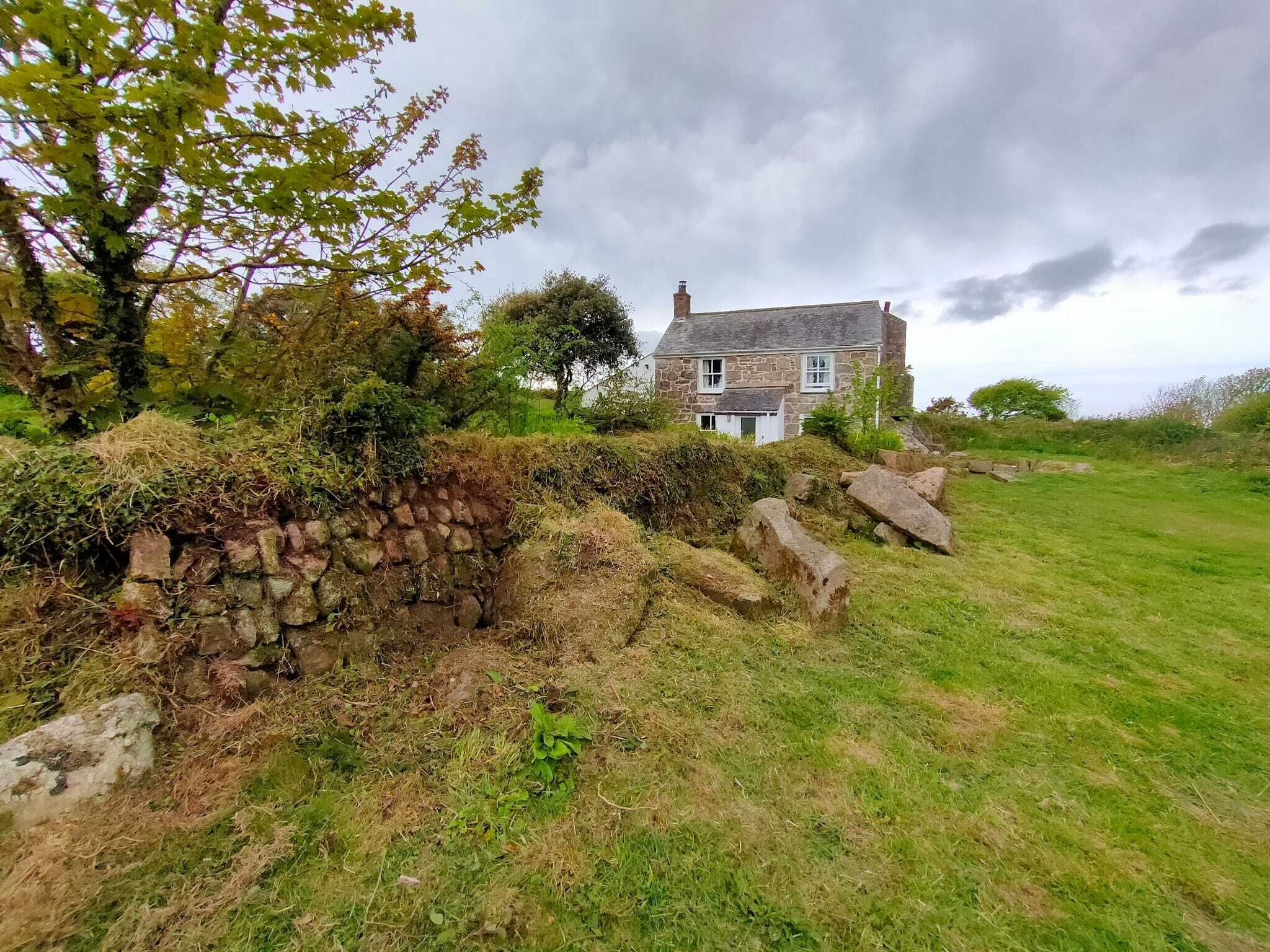 A stone house with a sloped roof, surrounded by a grassy field and a stone wall.