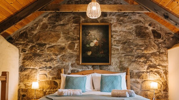 A bedroom with a stone wall, wooden ceiling beams, a bed with a wooden headboard, two bedside lamps, and a hanging light fixture. - Remote Cottage with Woodburner Surrounded by Farmland  (Redruth)