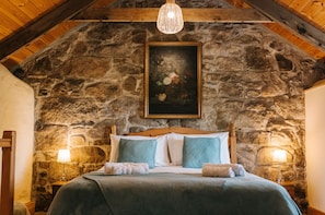 A bedroom with a stone wall, wooden ceiling beams, a bed with a wooden headboard, two bedside lamps, and a hanging light fixture.