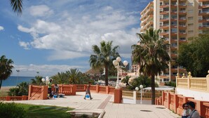 A coastal promenade with a palm-lined walkway, a beach in the background, and a multi-story building with balconies.