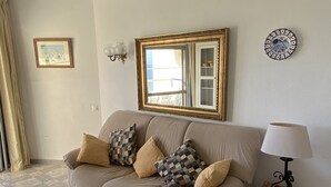 A living room with a beige sofa, patterned cushions, a small table with a bowl, a mirror with a decorative frame, a wall-mounted lamp, and a framed picture.