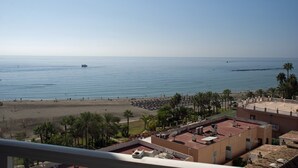 A coastal view with a beach, palm trees, and a building with a flat roof and satellite dishes.