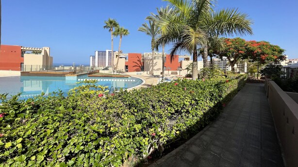 A pool area with a clear view of the ocean, surrounded by palm trees and modern buildings. - Spacious 2 bedroom apartment, large terrace, sea view, Paraiso II complex (Adeje)
