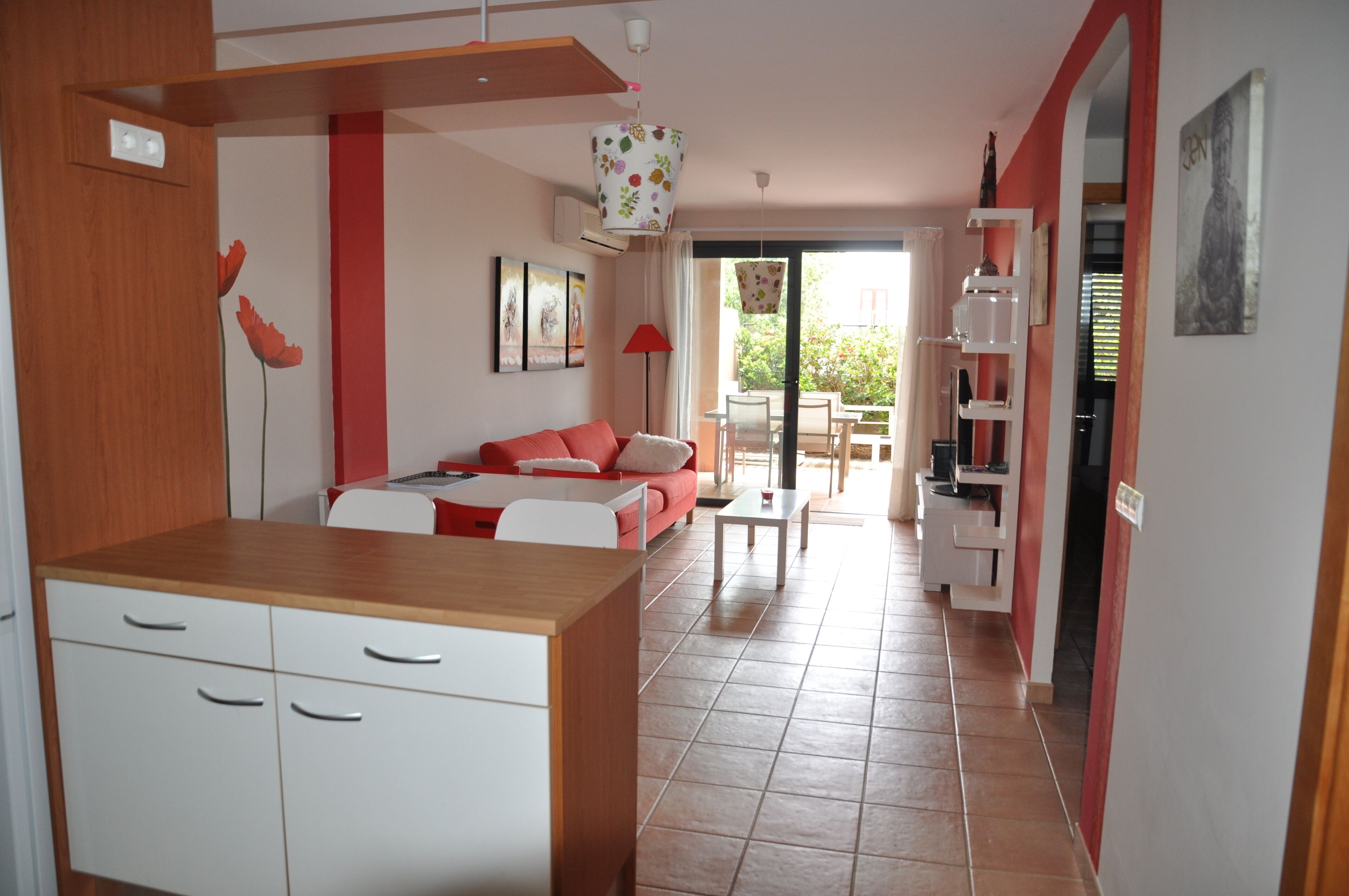 Una cocina moderna con armarios blancos y encimera de madera, un sofá rojo con una mesa blanca, y un comedor con una lámpara roja.