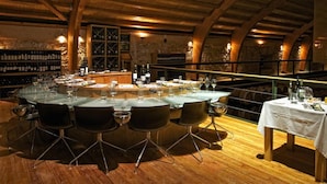 Un restaurante con una gran mesa redonda preparada para comer, rodeada de sillas. Se observan estanterías con vino al fondo y una zona de bar con botellas.
