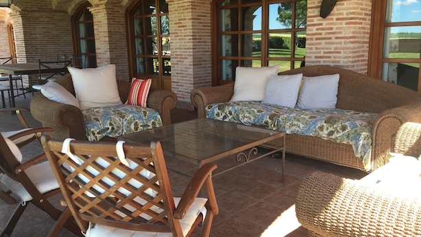 Un patio cubierto con muebles de mimbre, una mesa de vidrio y paredes de ladrillo.