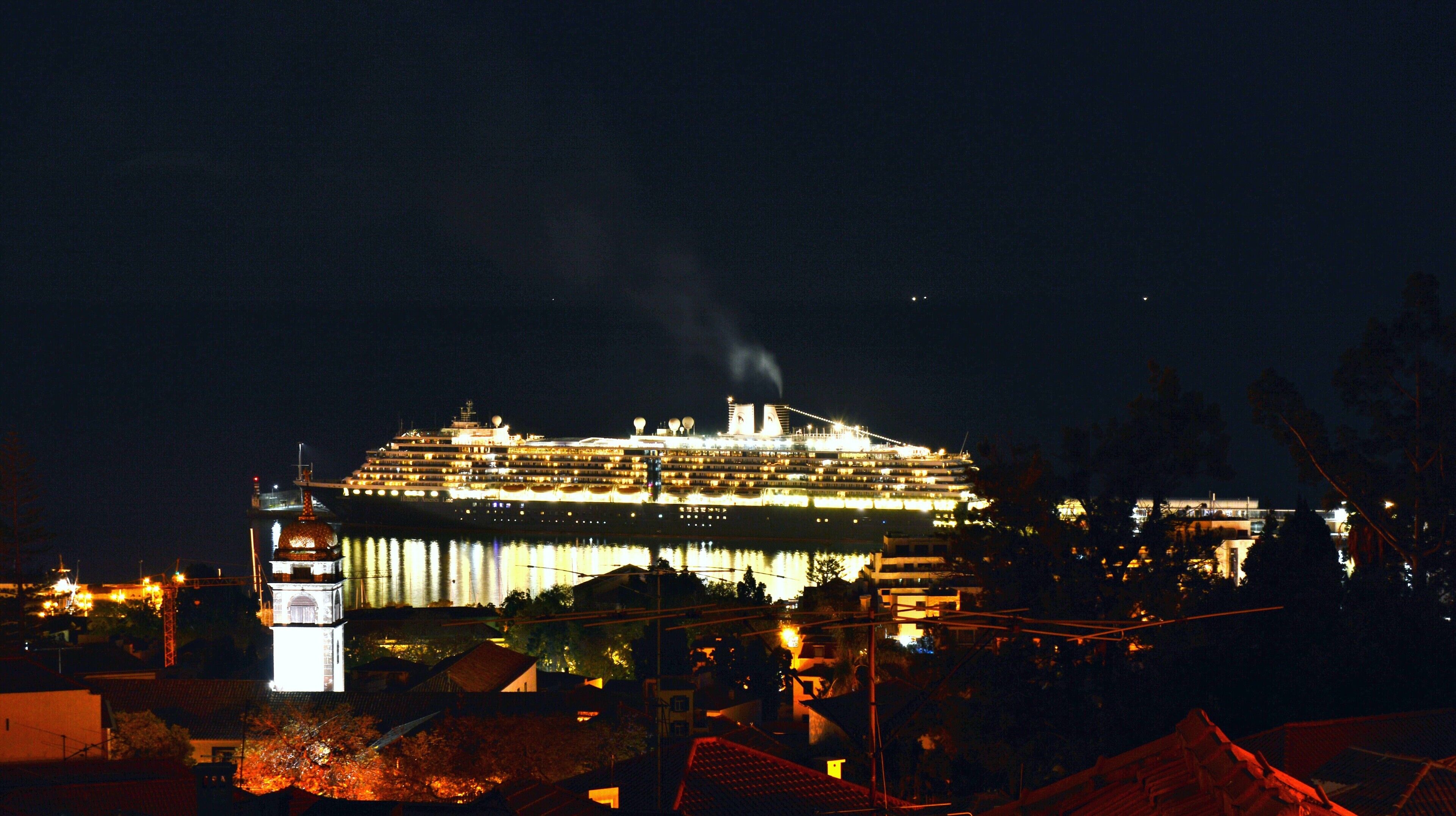 A large cruise ship docked at a port with multiple decks and a prominent smokestack emitting smoke.