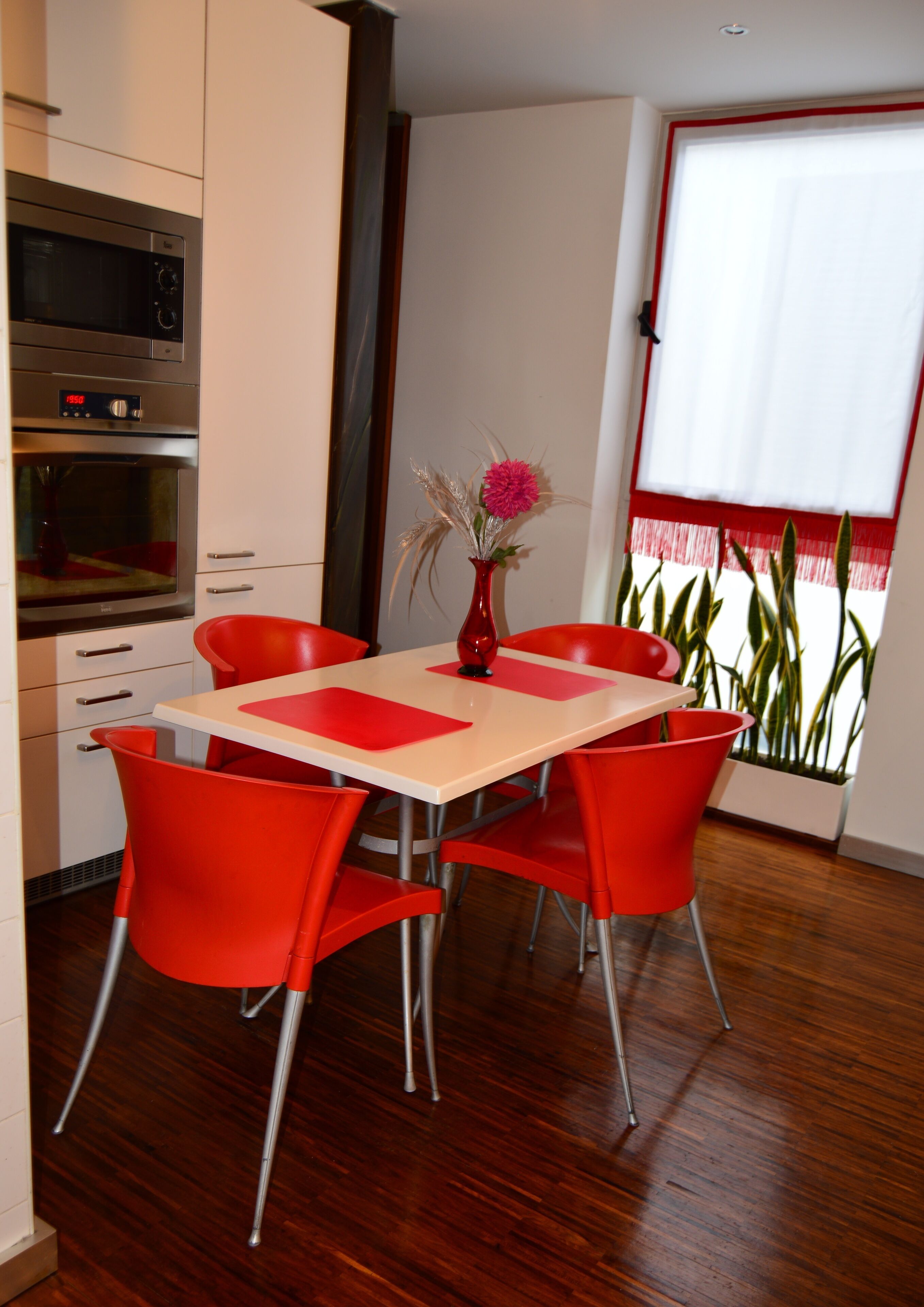 A modern kitchen with a built-in microwave and oven, a dining table with red chairs, and a window with red curtains.