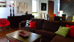 Ein modernes Wohnzimmer mit braunem Sofa, roten und grünen Kissen, einem Holztisch und einem roten Sessel. Zwei Wandbilder und ein großes Fenster sorgen für natürliches Licht.