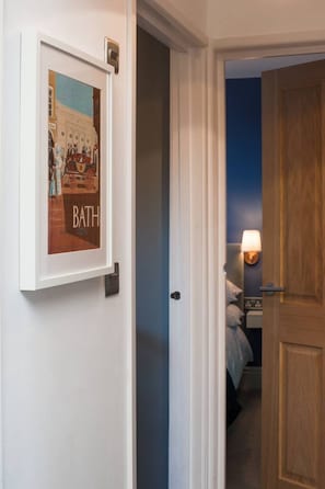 Un couloir orné d’un tableau encadré représentant une scène de bain, une porte donnant sur une chambre avec un lit et une table de chevet, et un luminaire fixé au mur.