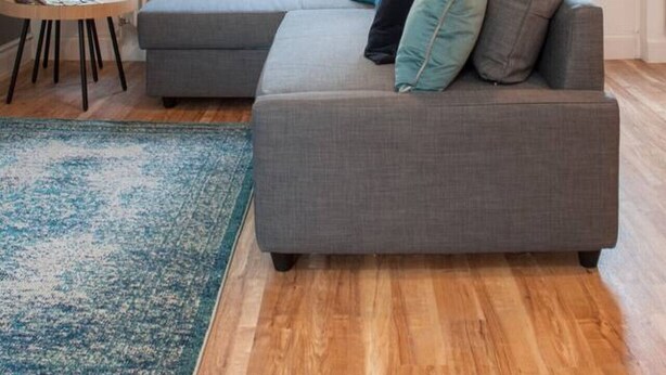 Ein modernes Wohnzimmer mit einem grauen Ecksofa, einem runden Holztisch mit einer Pflanze und einem blauen Teppich.