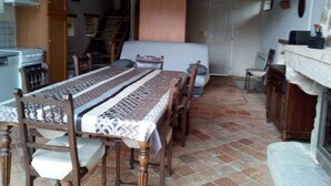 Un comedor con una larga mesa de madera, sillas y mantel estampado. Se ve un sofá y una pequeña mesa con un televisor al fondo.