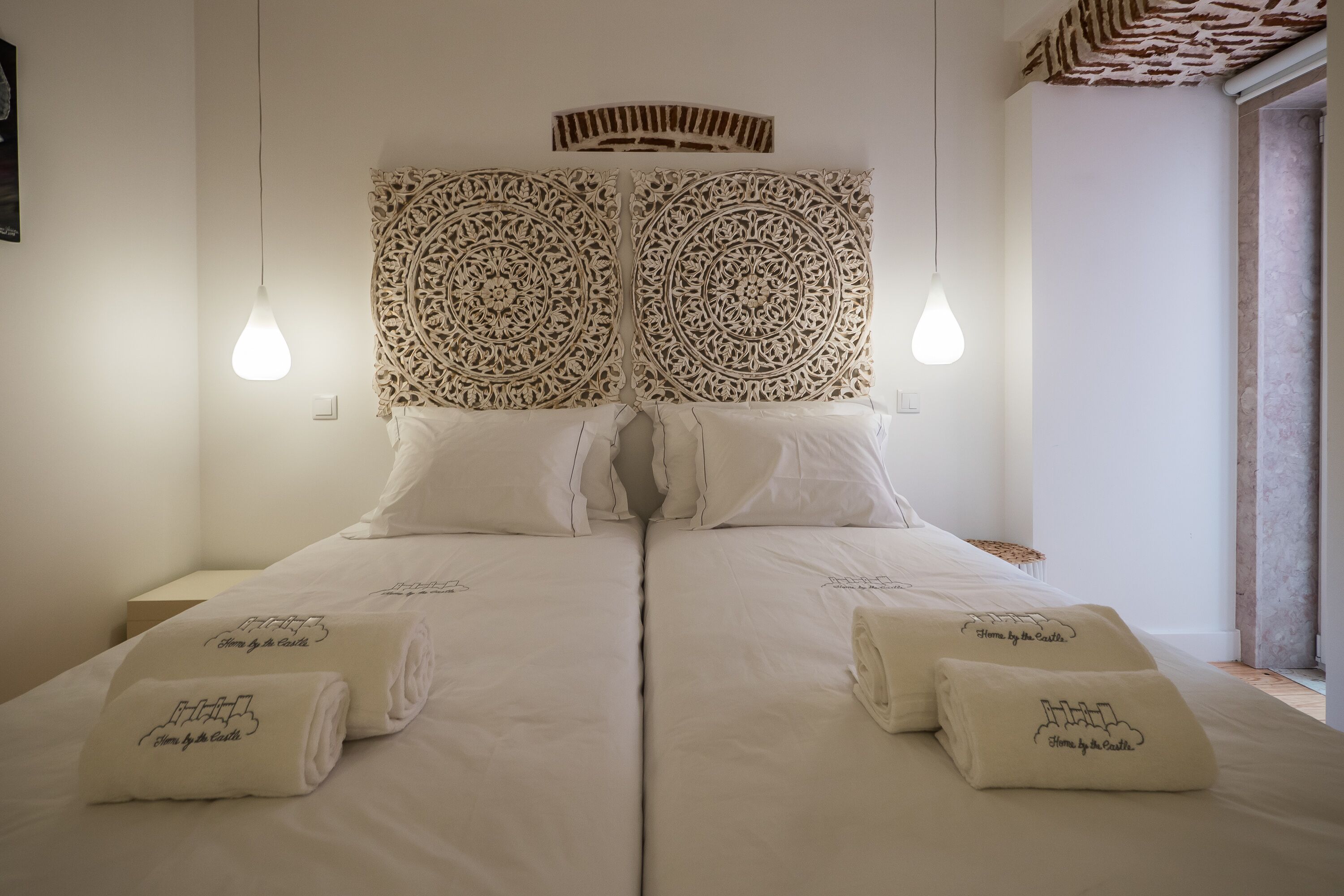 Una cama doble con ropa blanca y dos toallas, un cabecero decorativo y dos lámparas colgantes.
