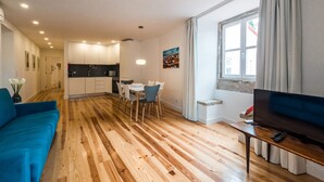 A modern living room with a blue sofa, wooden flooring, a dining area with a table and chairs, a kitchen in the background, and a TV on a wooden stand.