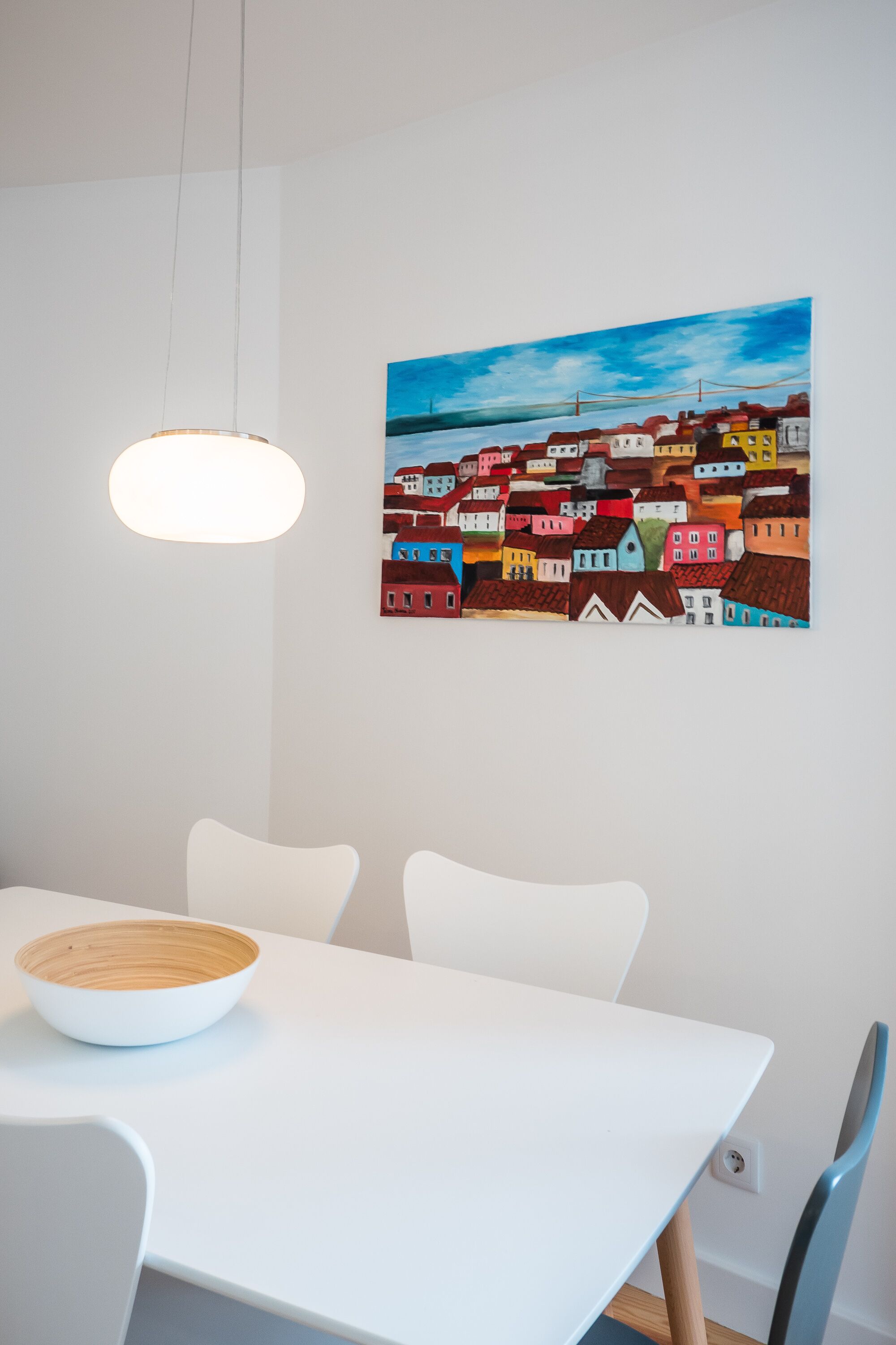 Ein Essbereich mit einem weißen Tisch, einer Schüssel und einem Bild von bunten Häusern an der Wand.