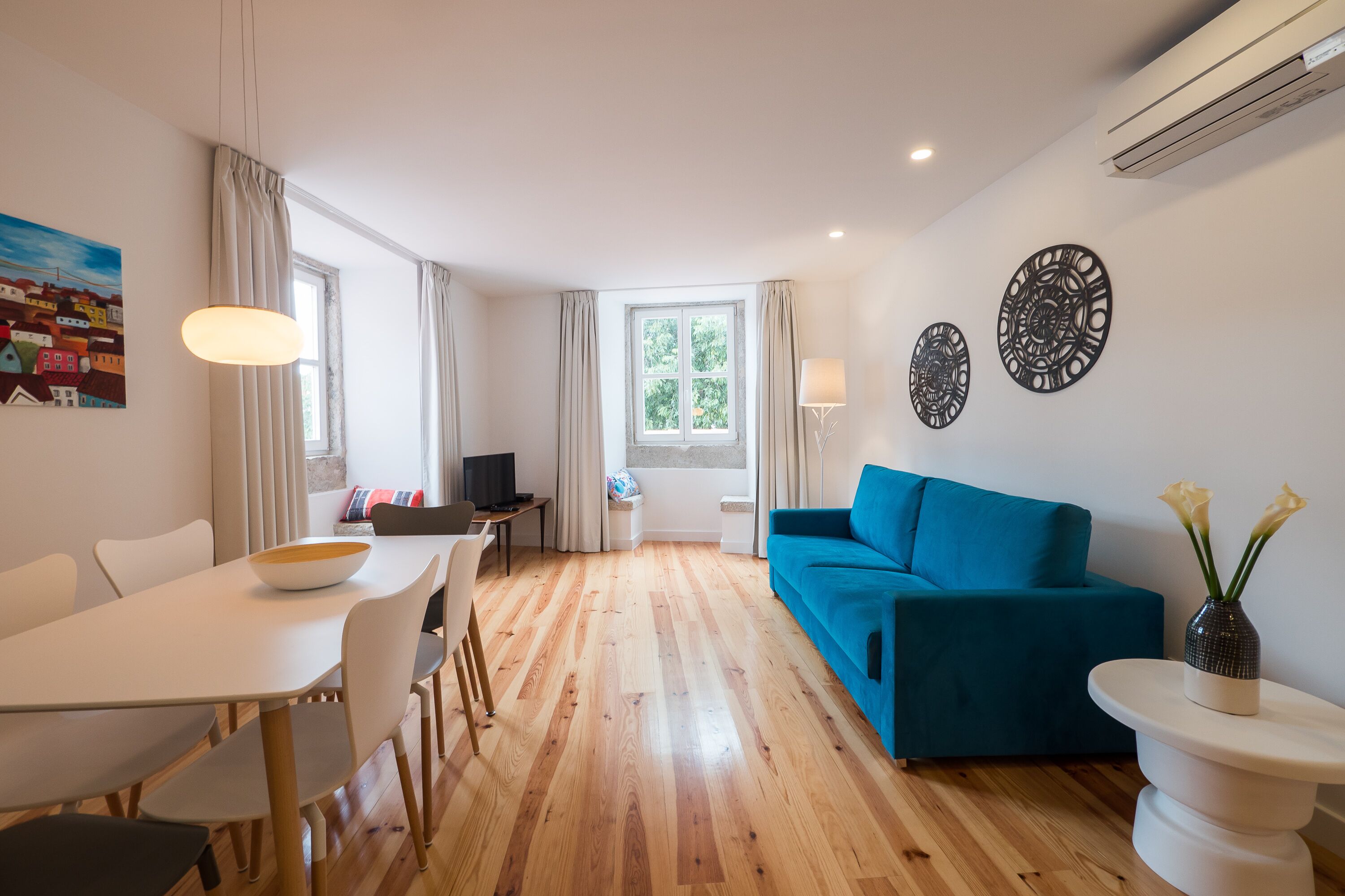 Un salon moderne avec un canapé bleu, une table à manger avec des chaises, un vase blanc rempli de fleurs et un tableau accroché au mur.