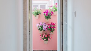 A narrow hallway with a wooden floor, a pink wall, and a sliding door leading to an outdoor area with hanging flower planters.