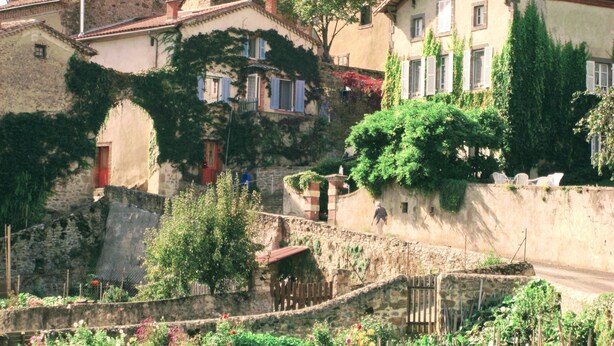 A traditional European village with stone buildings, some with ivy, and a stone wall with a gate.