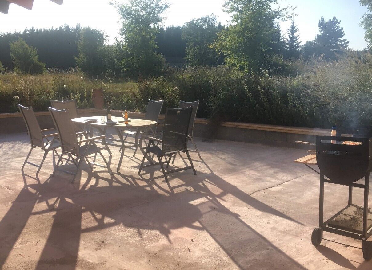 Une terrasse extérieure avec une table ronde et des chaises, un barbecue et de la verdure tout autour.