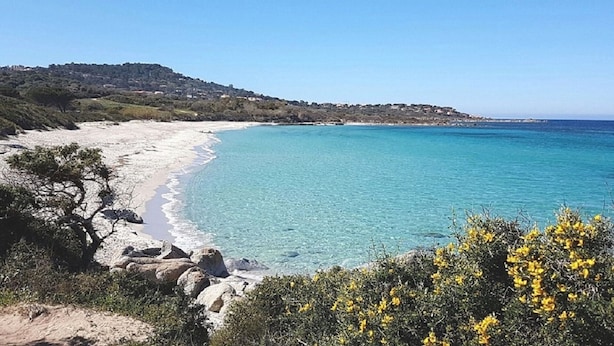 Un paysage côtier avec une plage de sable, des eaux d’un bleu limpide et une chaîne de collines en arrière-plan.
