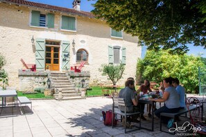 A stone building with a wooden door and green shutters, a staircase leading to the entrance, and a patio area with outdoor seating.