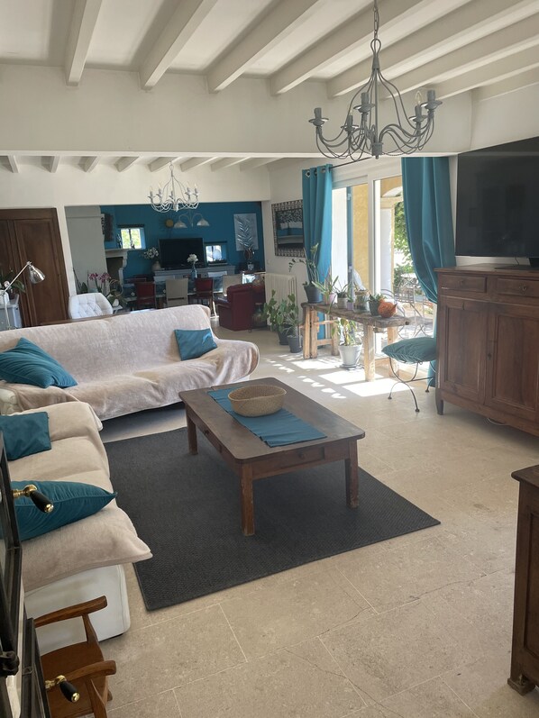 A spacious living room with a sofa, coffee table, and a TV. There is a kitchen area in the background with a dining table and chairs.