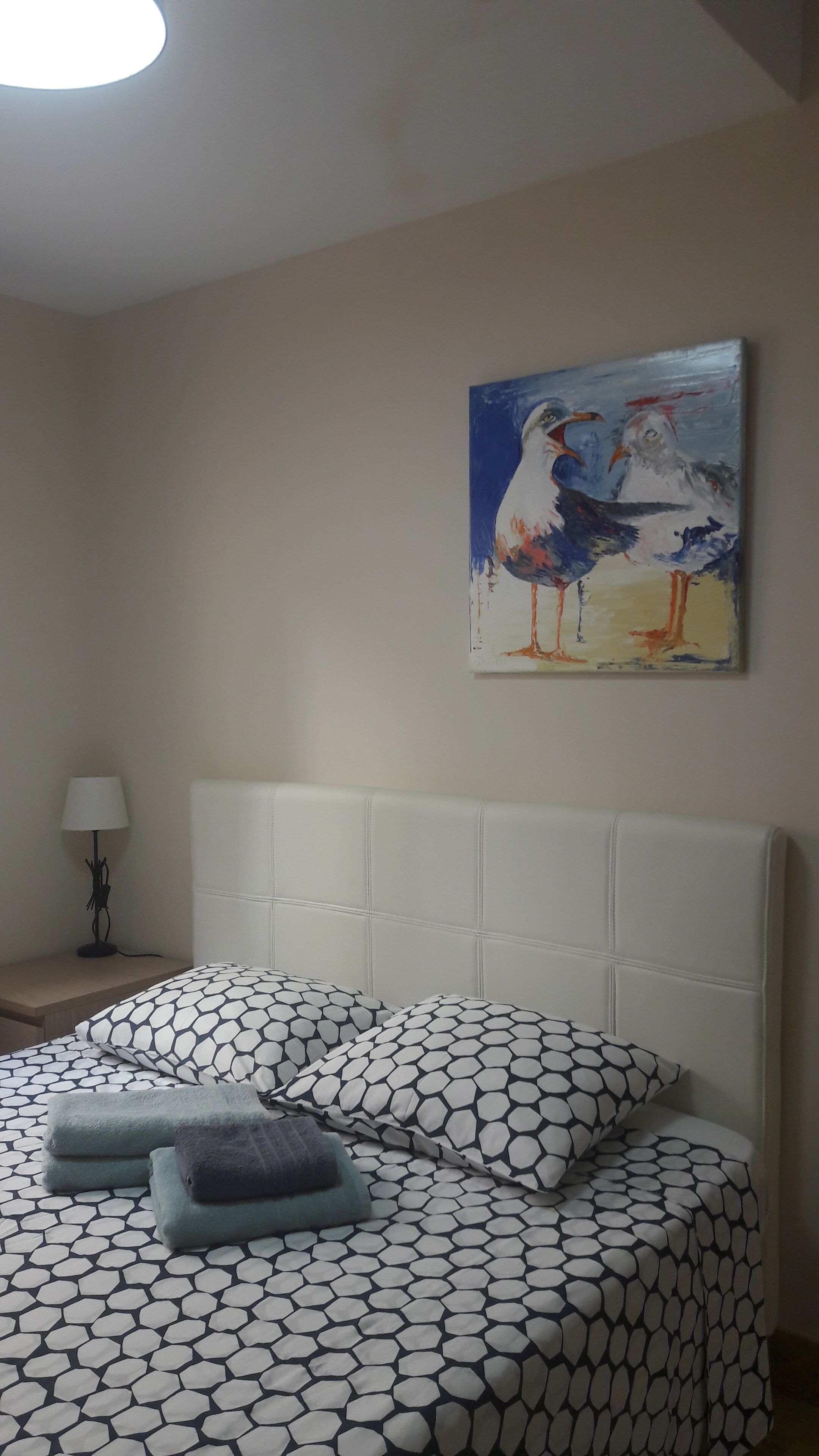 Ein Schlafzimmer mit einem Bett, das eine geometrische Tagesdecke, einen weißen Kopfteil und ein Vogelbild an der Wand zeigt.