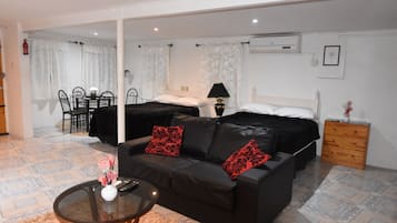 A hotel room with a black leather sofa, a small round table with a vase, a bed with black bedding and red pillows, a wooden nightstand, and a dining area with a table and chairs.