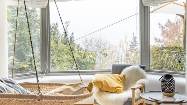Ein gemütliches Zimmer mit Hängestuhl, Sofa, einem kleinen Tisch und großen Fenstern mit Blick auf grüne Natur.