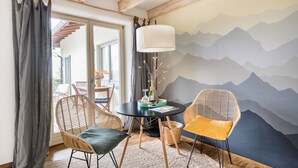 Ein gemütlicher Raum mit einem runden Tisch, Korbstühlen, einer Lampe und einem Wandbild von Bergen.