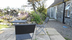 Ein Außenbereich mit Steintisch und zwei Stühlen, umgeben von einem Garten mit Steinen und blühenden Pflanzen.