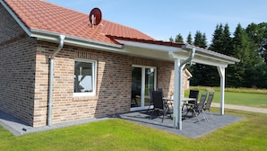 Une maison en briques avec un toit recouvert de tuiles rouges, un patio abrité avec une table et des chaises, et une grande fenêtre.