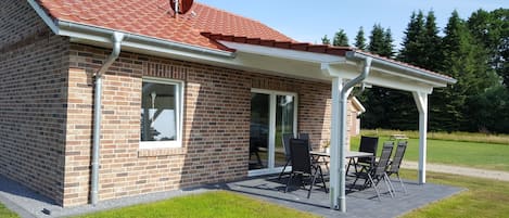 Et murhus med rødt tegltag, en overdækket terrasse med et bord og stole, og et stort vindue.