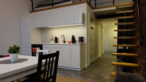 Een moderne keuken met witte kasten, een zwarte waterkoker en een koffiezetapparaat. Er staat een eettafel met stoelen en een bank. Een trap met houten treden leidt naar een andere kamer.