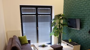Een moderne woonkamer met een bank, voetenbankje en een kleine tafel. Er is een groot raam met zonwering, een groene plant en een televisie die aan een bakstenen muur is bevestigd.