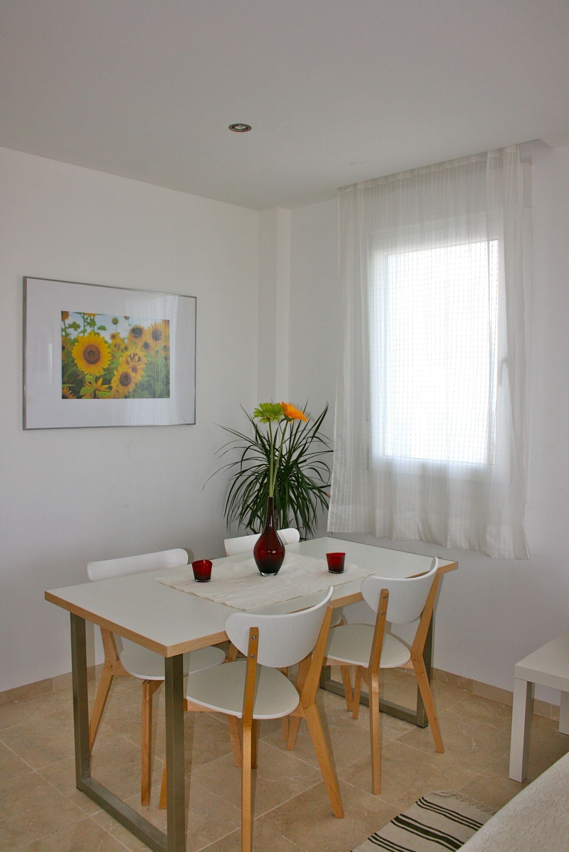A dining area with a white table and chairs, a vase with flowers, a framed picture on the wall, and a window with sheer curtains.