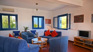 Ein Wohnzimmer mit blauer Sofagarnitur, einem runden Couchtisch und einem Fernseher auf einem Holzständer. Zwei Fenster mit blauen Rahmen sind vorhanden, und an den Wänden befindet sich Kunstwerk.