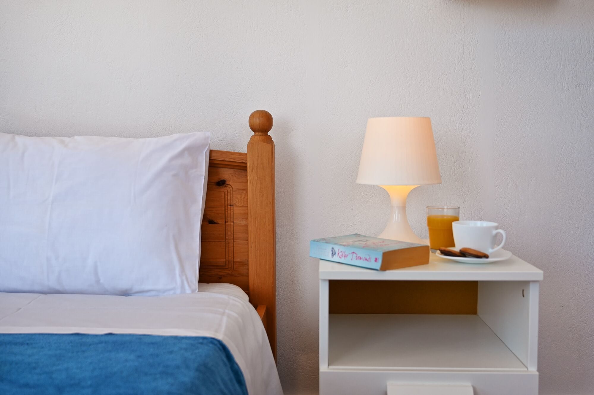 A neatly made bed with a white pillow and a blue blanket, a wooden headboard, a white nightstand with a lamp, a book, a glass of orange juice, and a cup.