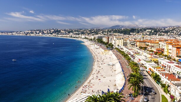 A coastal cityscape with a wide beach, clear blue waters, and a dense urban area with buildings and palm trees.