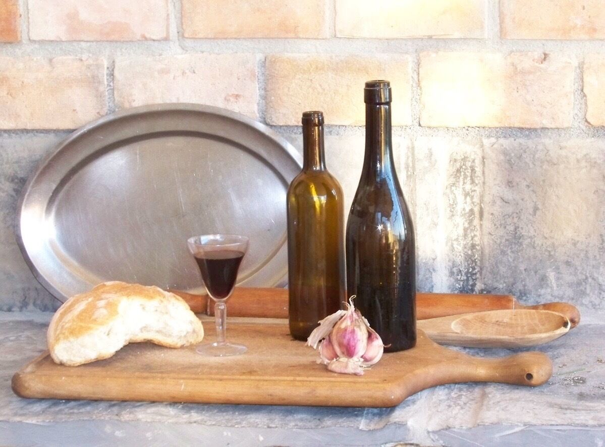 Tagliere in legno con una pagnotta di pane, un bicchiere di vino rosso, due bottiglie e un bulbo d'aglio.