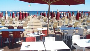 A beachside restaurant with outdoor seating, red umbrellas, and a view of the sea.