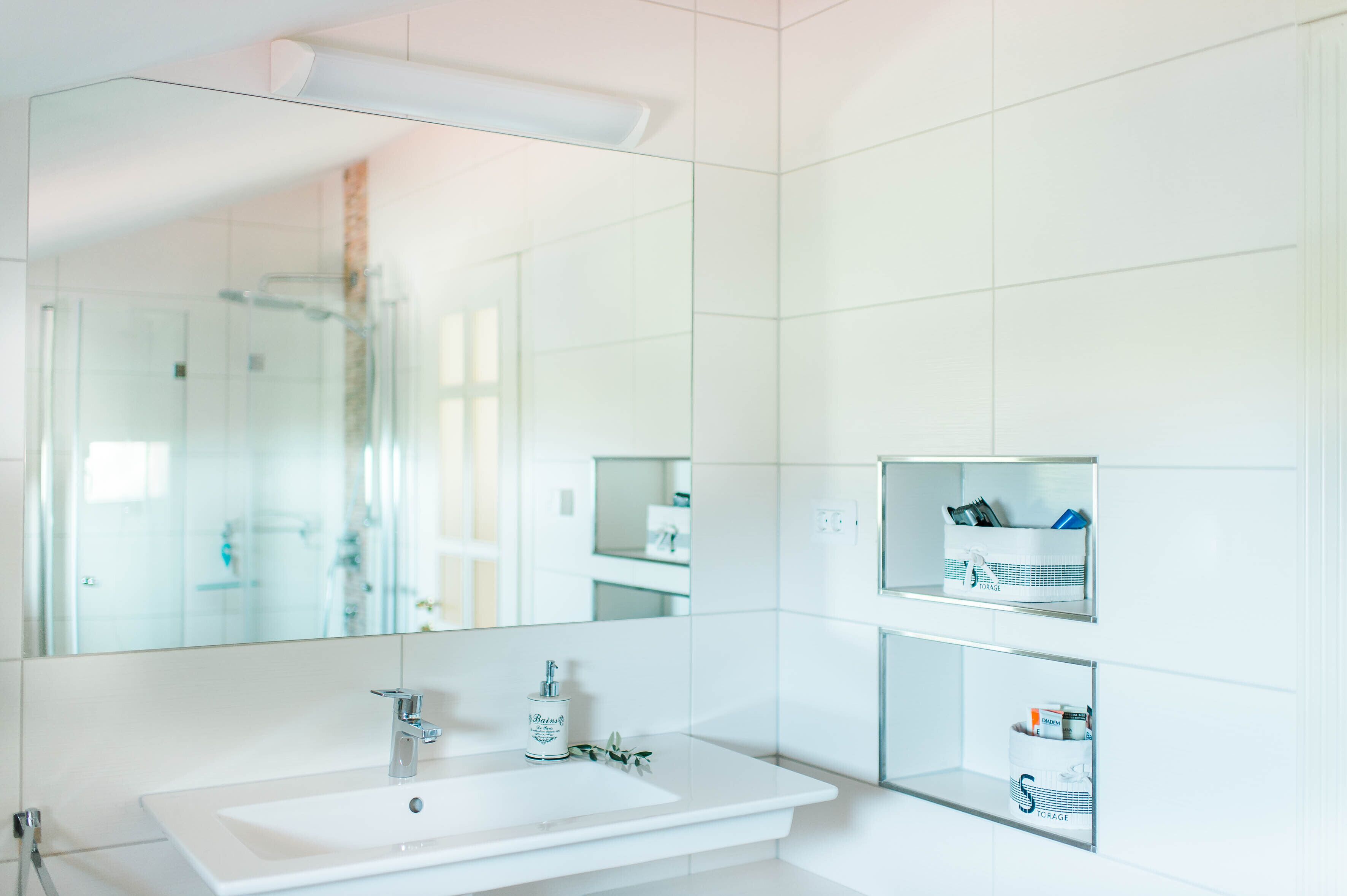 Une salle de bain moderne avec un grand miroir, un lavabo blanc et des étagères intégrées pour le rangement.
