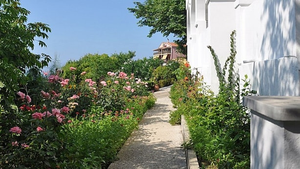 Uma trilha ladeada por vegetação exuberante e flores cor-de-rosa que leva a um edifício branco com arcos.